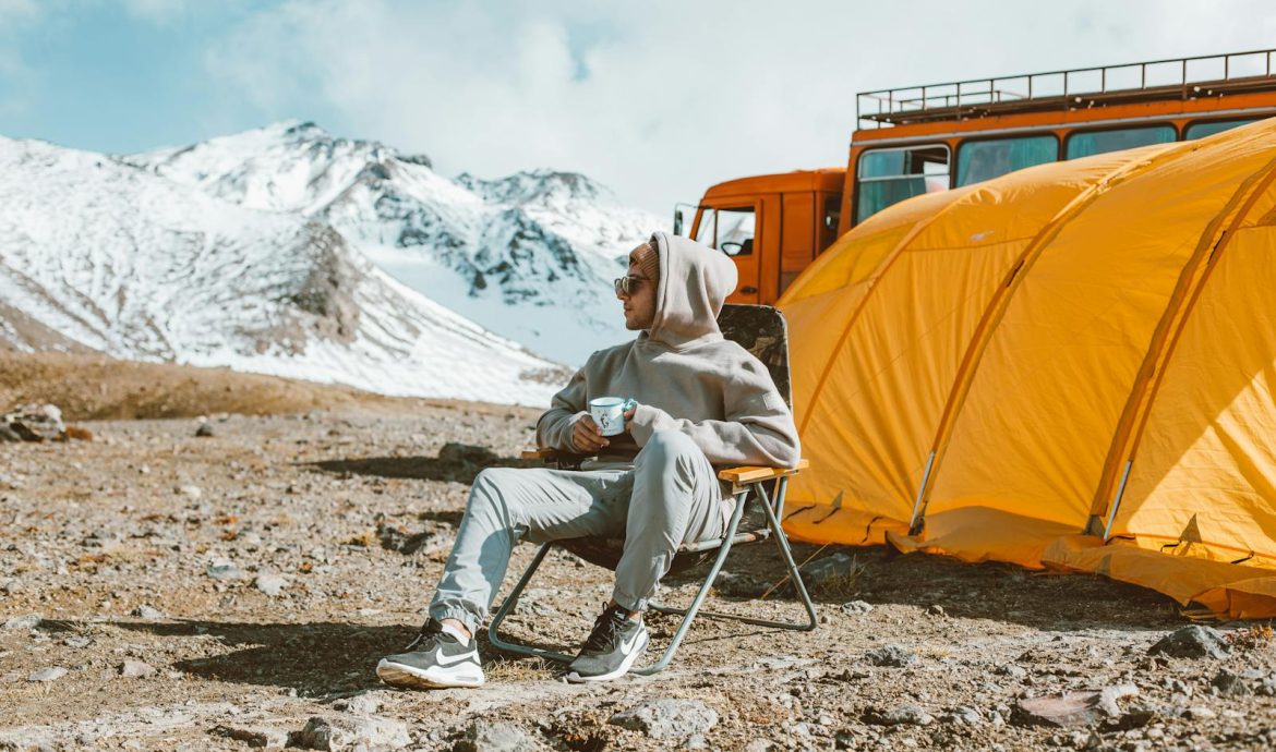 Full length of male camper sitting on folding chair next to tent in middle of mountain valley and enjoying views